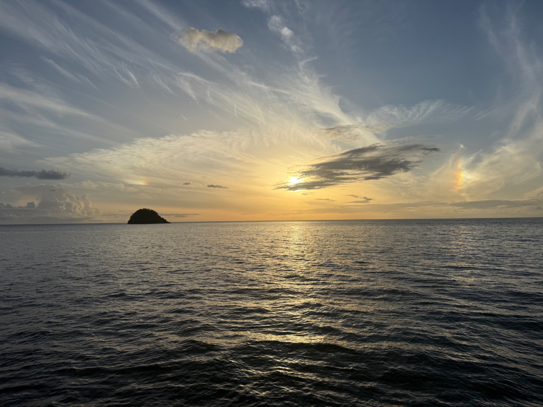 A golden sunset over calm water with a small island silhouette and sunbows in the clouds