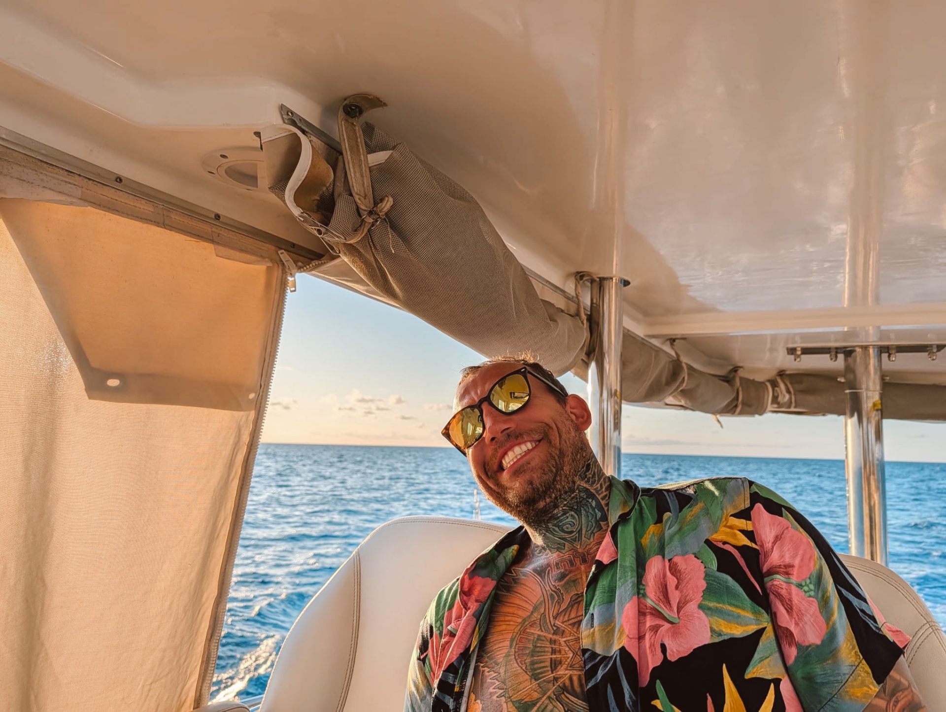 Cody smiling under the boat's canopy in sunglasses and an open floral shirt