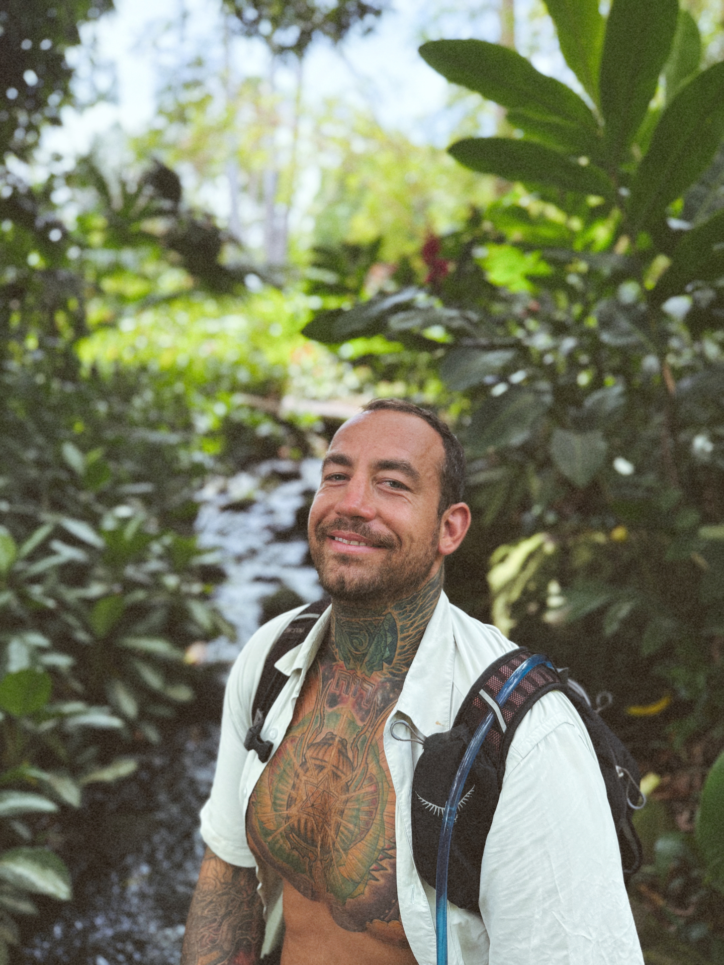 Cody smiling on a jungle hike, hydration pack on, a rocky creek behind him