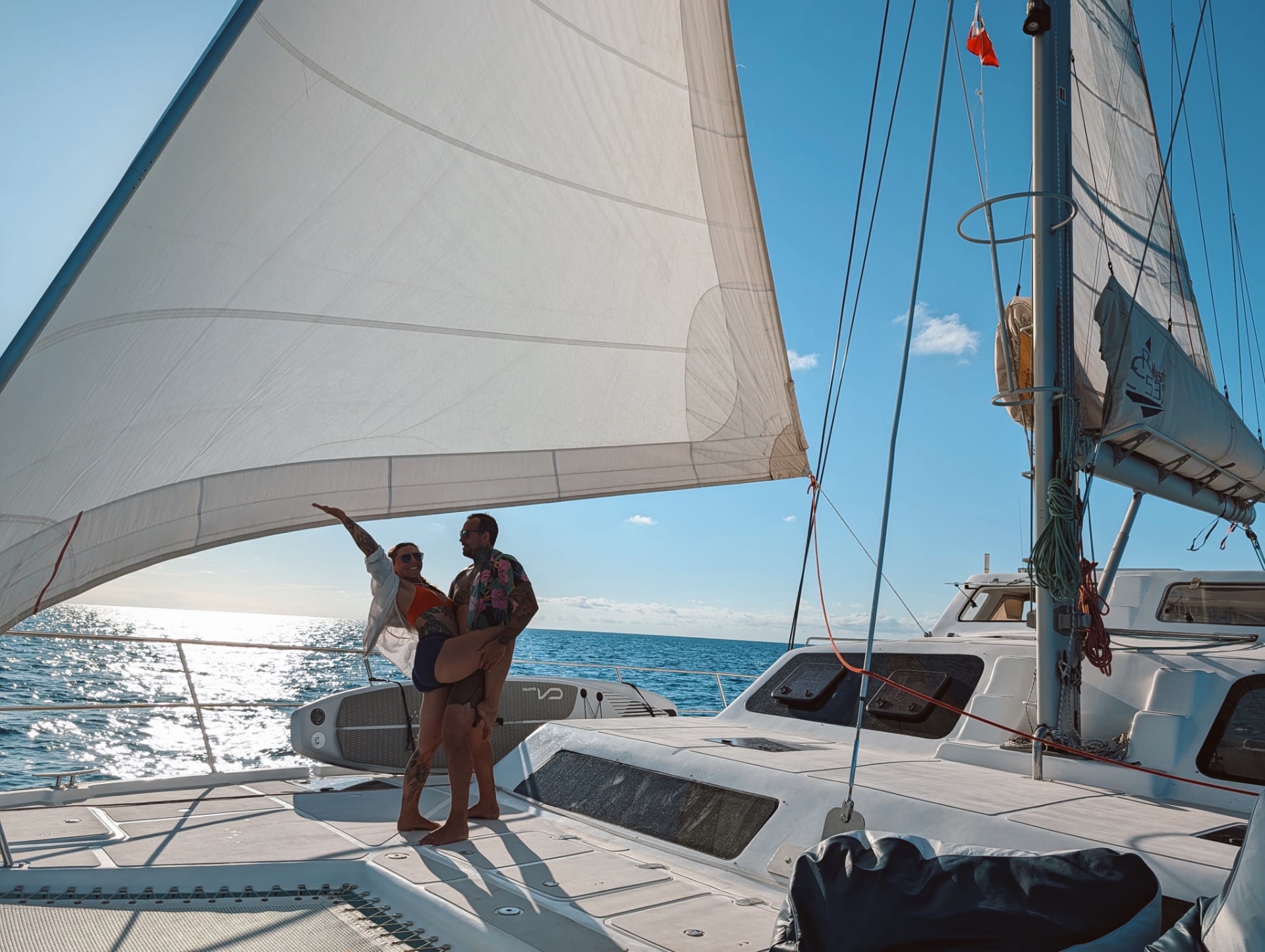 Gina and Cody posing together on deck beneath the billowing white sail of the catamaran