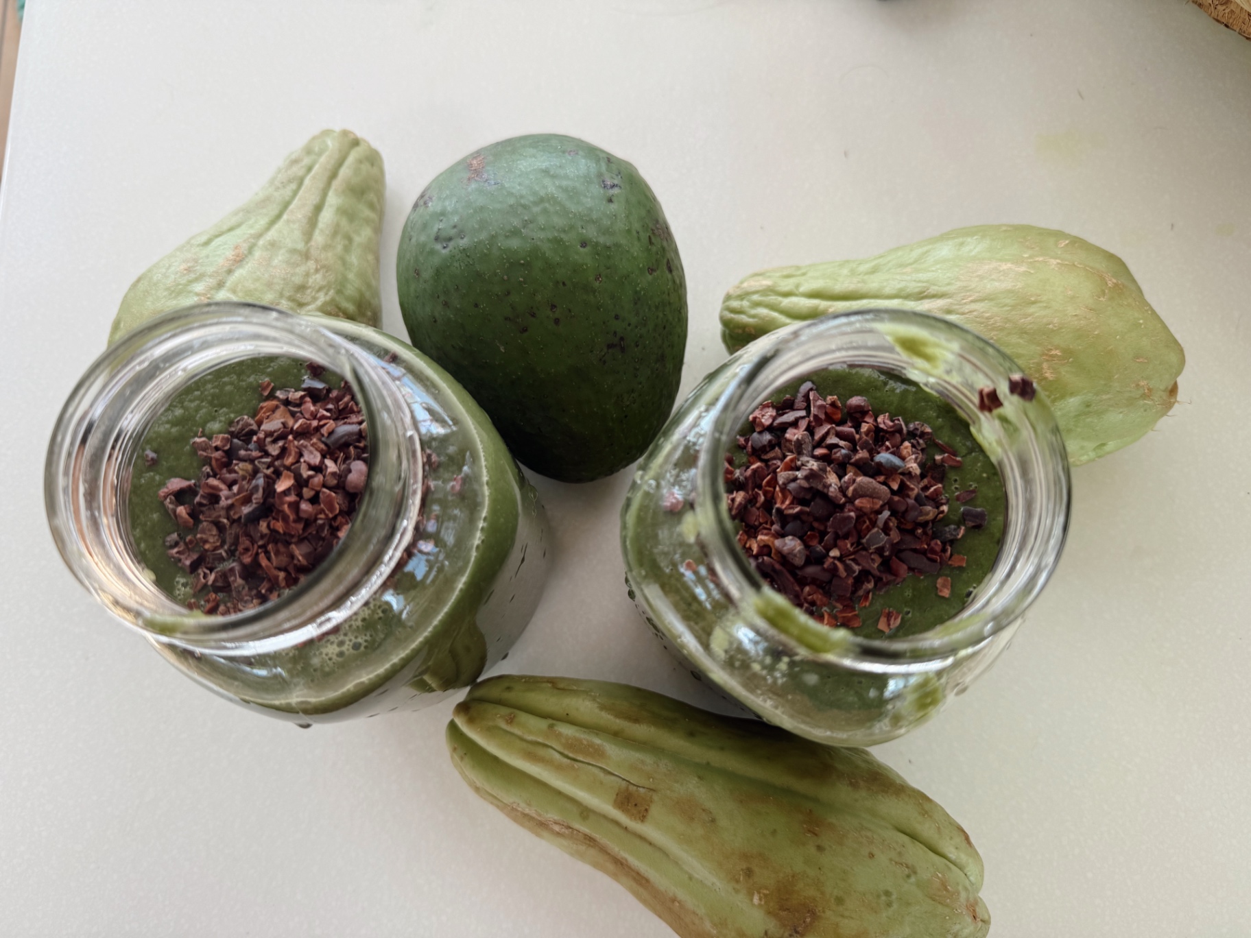 Two jars of green smoothie topped with cacao nibs, surrounded by avocado and christophene