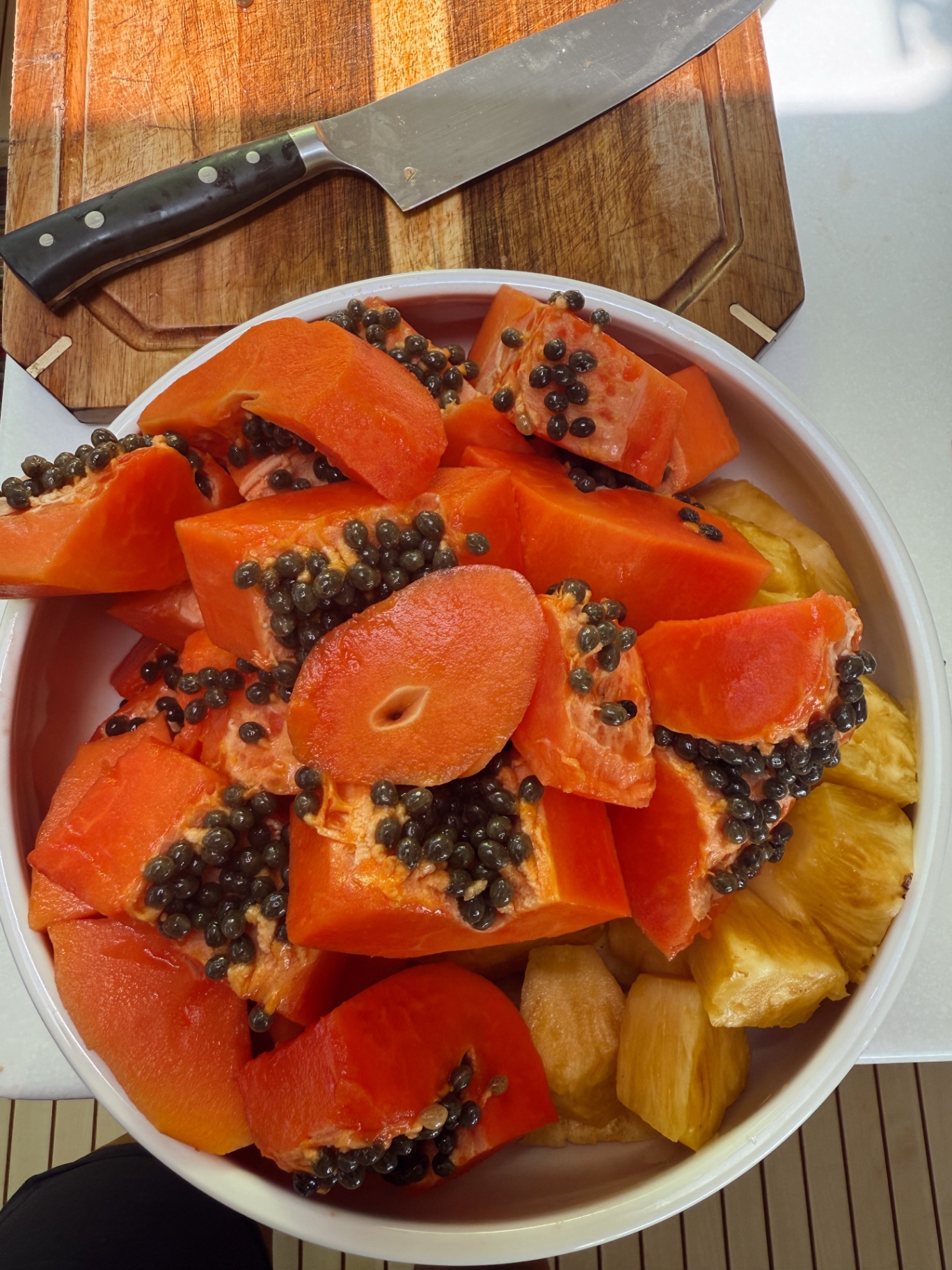 A white bowl of sliced papaya and pineapple on a wooden board, a knife resting behind it